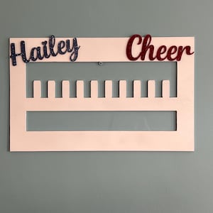以下が含まれることがあります： 青いグリッターで「Hailey」、赤いグリッターで「Cheer」と書かれた白い木製のチアリーディングディスプレイボード。ボードには、アイテムを表示するための複数のスロットがあります。