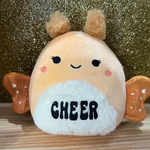 May include: A plush, orange-colored toy with a smiling face, small brown antennae, and brown wings. The toy has a white fluffy belly with the word "CHEER" in black letters. The toy is on a wooden shelf with a gold glitter background.