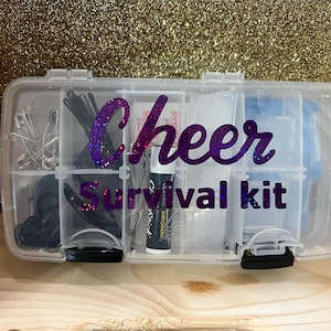 Puede incluir: Un organizador de plástico transparente con la etiqueta "Cheer Survival Kit" con letras de purpurina moradas. El kit contiene varios artículos, incluyendo imperdibles, horquillas y un tubo de bálsamo labial. El fondo es una superficie dorada con purpurina.