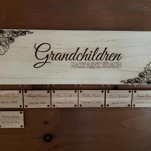 May include: Wooden sign with the words "Grandchildren MAKE LIFE GRAND" in a cursive font. The sign has floral designs in the corners and includes name tags with birthdates. A decorative piece for a family.