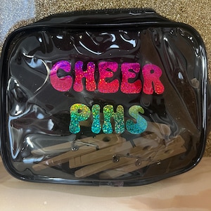 Può includere: Un astuccio nero e trasparente con cerniera con le parole "CHEER PINS" in lettere colorate e glitterate. All'interno ci sono diverse mollette di legno. Lo sfondo è dorato e scintillante.