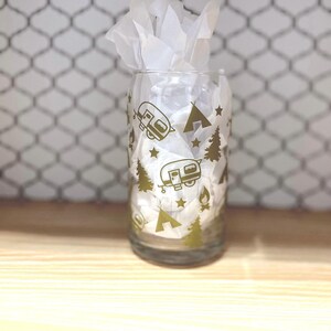 May include: A clear glass with a gold design of camping trailers, stars, and pine trees. The glass is partially filled with white tissue paper.