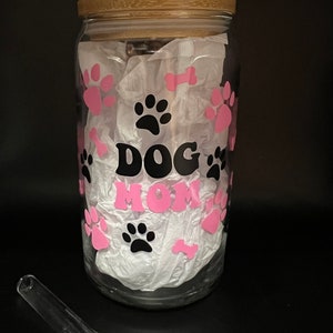 May include: A clear glass jar with a bamboo lid and a pink and black "Dog Mom" design. The jar is filled with white tissue paper and has pink paw prints and bone designs. A clear glass straw is included.