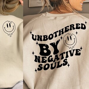 Può includere: Una felpa beige con una stampa grafica nera che dice "Unbothered by Negative Souls" con un'emoticon sorridente che cola.