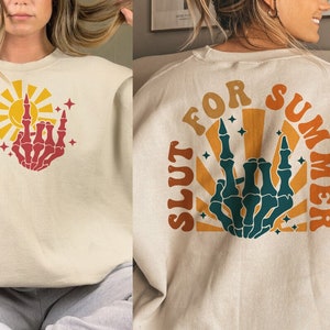 May include: Cream-colored sweatshirt with a retro design featuring a red skeleton hand giving the middle finger, a yellow sun, and the text "FOR SUMMER SLUT" on the back. The design includes orange and teal accents.