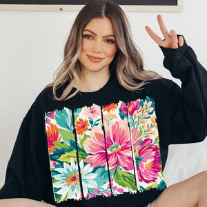May include: Black sweatshirt with a colorful floral print design. The print features a variety of flowers in shades of pink, orange, yellow, green, and blue. The flowers are arranged in a vertical pattern and are surrounded by a white background.
