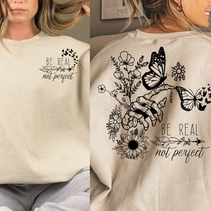 May include: A beige sweatshirt with a black graphic print of a hand holding flowers and butterflies. The text "Be real not perfect" is printed on the sweatshirt.