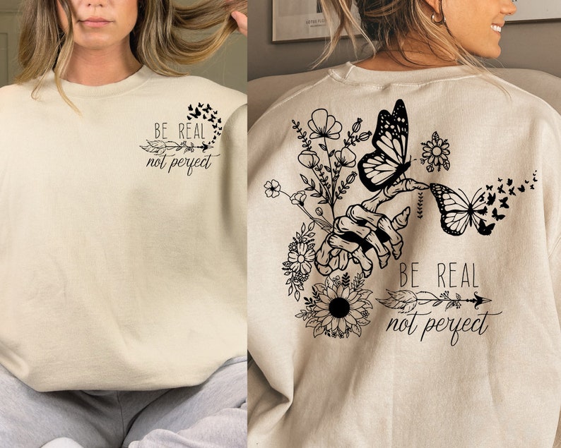 May include: A beige sweatshirt with a black graphic print of a hand holding flowers and butterflies. The text "Be real not perfect" is printed on the sweatshirt.