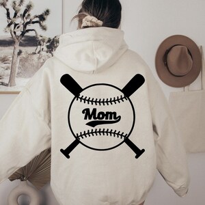 May include: A white hoodie with a black baseball graphic on the back. The graphic features a baseball with the word "Mom" in a vintage font and two crossed bats.