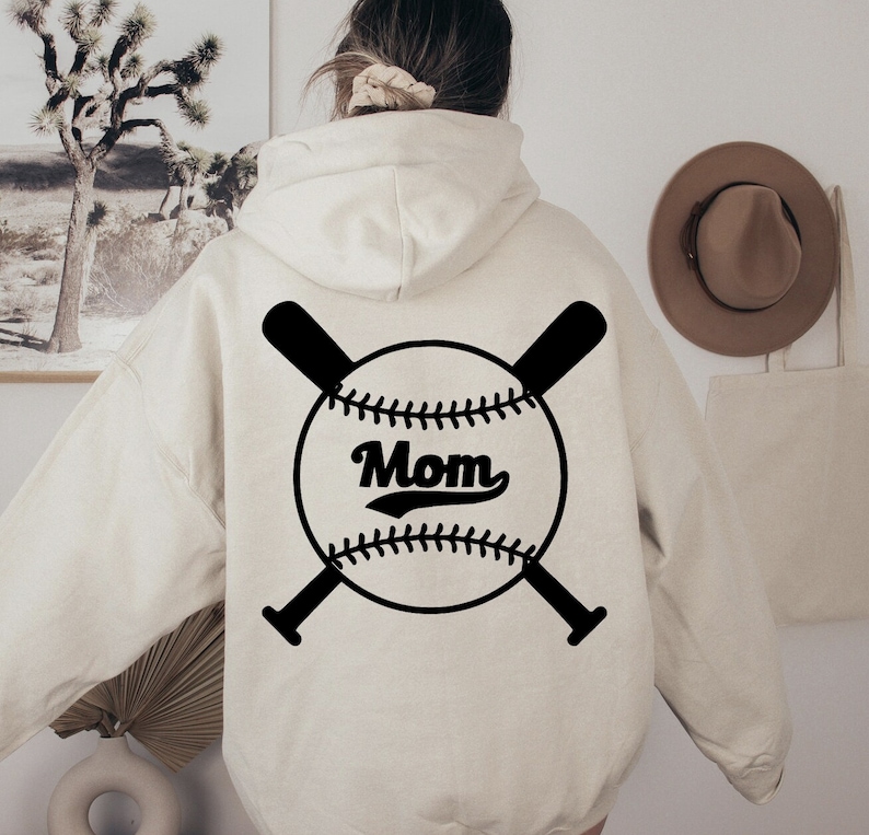 May include: A white hoodie with a black baseball graphic on the back. The graphic features a baseball with the word "Mom" in a vintage font and two crossed bats.