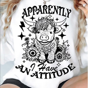 May include: White sweatshirt with a black and white illustration of a Highland cow with the text "Apparently I Have An Attitude" printed on it.