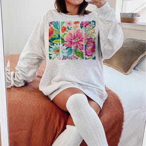 May include: A gray sweatshirt with a floral print design. The print features a variety of flowers in shades of pink, white, and green.