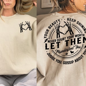 May include: A beige sweatshirt with a black graphic print on the back. The graphic features a circle with the text "Your Beauty. Keep Shining. Never Doubt Your Worth. Let Them Misunderstand You. Judge You. Gossip About You." inside. The graphic also features two skeleton hands making a heart shape.