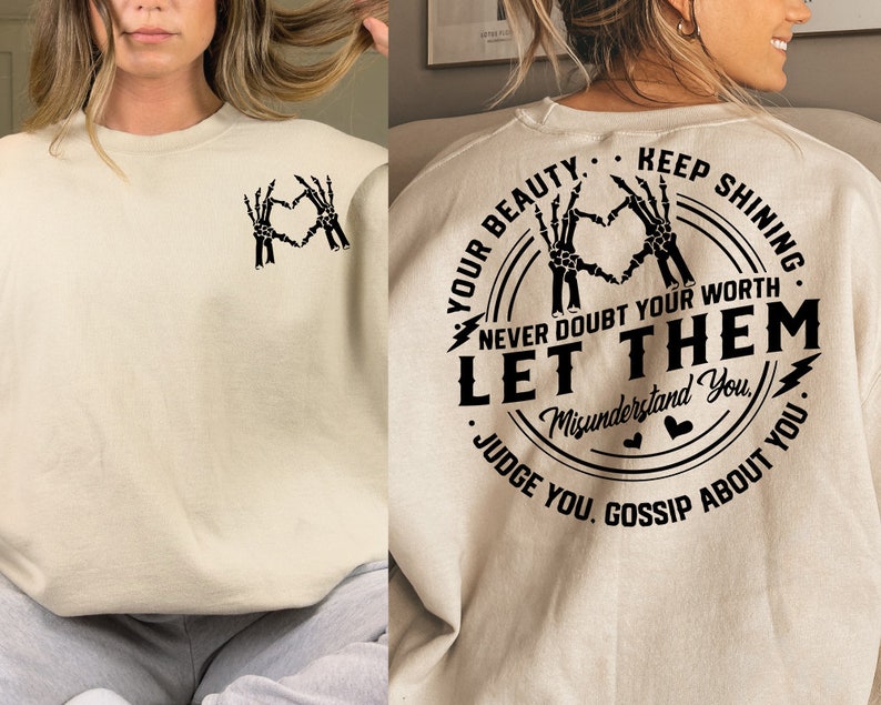 May include: A beige sweatshirt with a black graphic print on the back. The graphic features a circle with the text "Your Beauty. Keep Shining. Never Doubt Your Worth. Let Them Misunderstand You. Judge You. Gossip About You." inside. The graphic also features two skeleton hands making a heart shape.