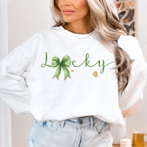 May include: White sweatshirt with the word "Lucky" in green script, featuring a green bow and gold coins. The long-sleeved top is a casual style, perfect for St. Patrick's Day or everyday wear.
