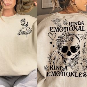 May include: A beige sweatshirt with black text and graphics. The text reads "Kinda Emotional Kinda Emotionless" and features a skull with flowers and a skeleton hand holding a rose.