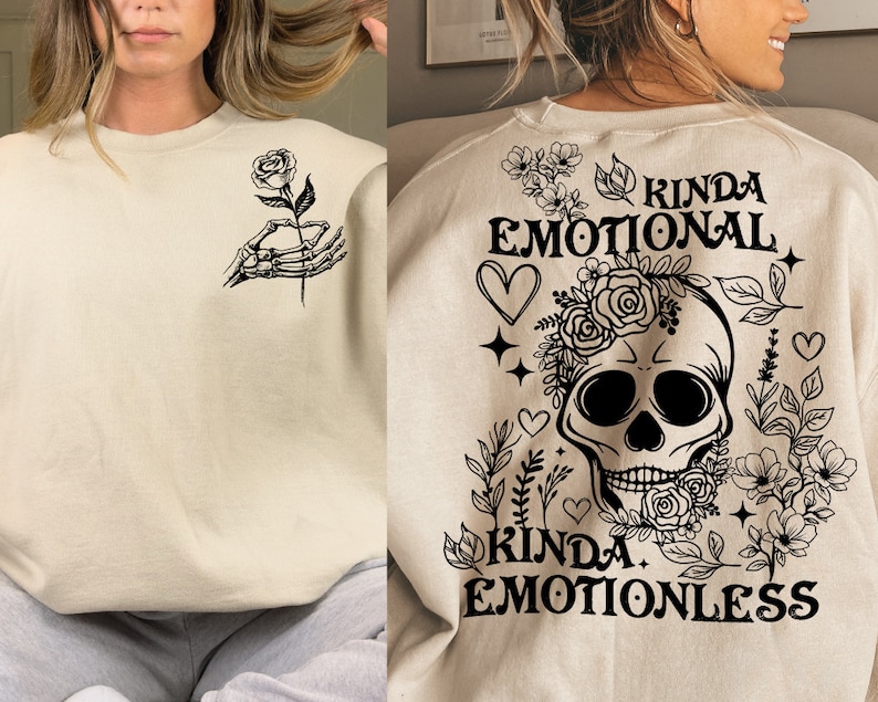 May include: A beige sweatshirt with black text and graphics. The text reads "Kinda Emotional Kinda Emotionless" and features a skull with flowers and a skeleton hand holding a rose.