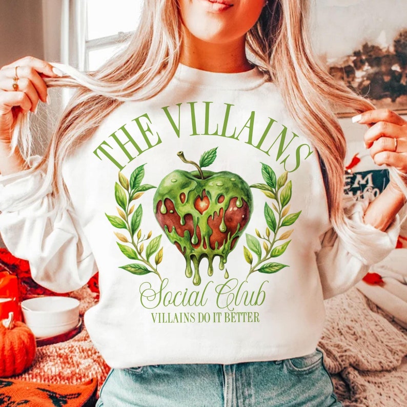 以下が含まれることがあります： 緑のリンゴのグラフィックと「The Villains Social Club」と「Villains Do It Better」のテキストが書かれた白いスウェットシャツ。