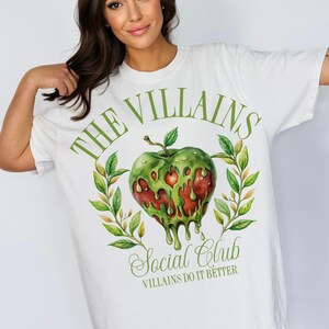 以下が含まれることがあります： 緑のリンゴのグラフィックと「The Villains Social Club」と「Villains Do It Better」のテキストが書かれた白いTシャツ。