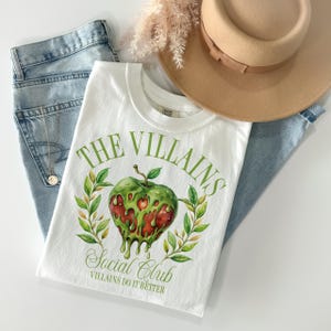 以下が含まれることがあります： 緑のリンゴのグラフィックが描かれた白いTシャツ。リンゴの中には赤いハートが滴り落ちています。リンゴの下には「The Villains Social Club」という文字が印刷されています。クラブ名の下には「Villains Do It Better」という文字が印刷されています。