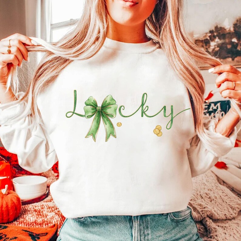 May include: White sweatshirt with the word "Lucky" in green script, featuring a green bow and gold coin accents. The design is a watercolor style, perfect for St. Patrick's Day or everyday wear.