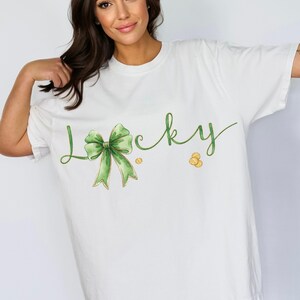 May include: White t-shirt with the word "Lucky" in green script, featuring a green bow and gold coins. The shirt is a loose fit, suitable for casual wear. The design has a watercolor effect, perfect for St. Patrick's Day.