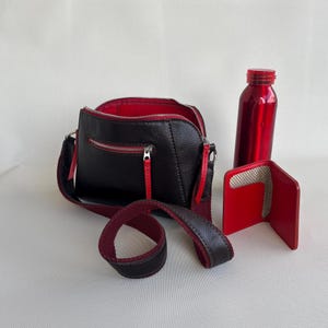 Puede incluir: Bolso bandolera de cuero negro con interior y cremalleras rojas, una correa a juego, una botella de agua roja y una cartera roja. El bolso tiene una cremallera curva y un pequeño bolsillo exterior.