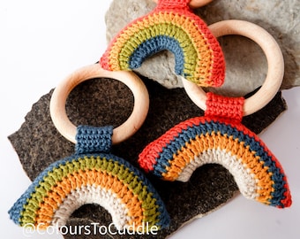May include: Three crocheted rainbow teethers attached to wooden rings. The rainbows are made with different colored yarn, including red, orange, yellow, green, blue, and beige. The rings are natural wood.