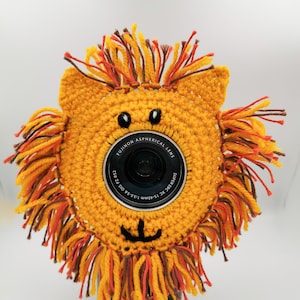 May include: A crocheted lion-shaped camera lens cover in yellow, orange, and brown. The cover has a lion's face with black eyes and a smiling mouth. The lens is visible in the centre. The text "@ColoursToCuddle" is at the bottom.