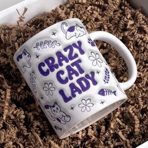 May include: White ceramic mug with a purple design that says "Crazy Cat Lady". The mug features a purple cat, flowers, and fish bones.