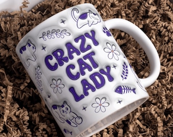 May include: White ceramic mug with a purple design that says "Crazy Cat Lady". The mug has a purple cat, flowers, and fish bones on it.