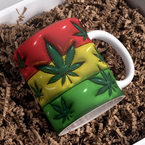 May include: A white ceramic mug with a red, yellow, and green design featuring stylized marijuana leaves. The mug is sitting in brown packing peanuts.