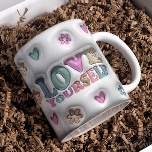 Puede incluir: Taza de cerámica blanca con un mensaje colorido "Love Yourself" y diseños florales.