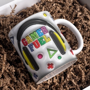 May include: White ceramic mug with a colorful gamer fuel design. The mug features a pair of headphones, a joystick, and other gaming symbols.