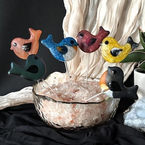 May include: A collection of six glass bird ornaments on clear stems, featuring various colors including orange, blue, dark red, yellow, dark green, and a black and orange bird, all arranged in a glass bowl filled with pink salt crystals.