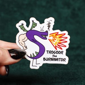 May include: A die-cut sticker featuring a purple dragon character with a muscular arm, green wings, and thin legs, breathing orange and yellow flames. The text "TROGDOR THE BURNINATOR" is printed in black capital letters next to the dragon illustration.
