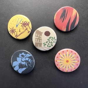 May include: Five circular pinback buttons featuring distinct graphic designs: a yellow button with palm trees and pyramids, a red and black abstract pattern, a beige button with a building and foliage, a dark blue bee illustration, and a pink starburst.