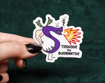 May include: A die-cut sticker featuring a purple dragon character shaped like the letter S with a muscular human arm, green wings, and thin legs. The dragon breathes orange and yellow flames, with the text TROGDOR THE BURNINATOR printed below.
