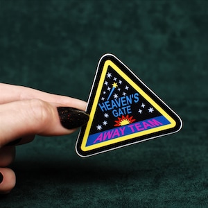 May include: A triangular sticker with a black background featuring a yellow border. The text reads HEAVEN'S GATE in blue and AWAY TEAM in pink. The design includes white stars, a blue shooting star, and a red and yellow sunburst graphic.