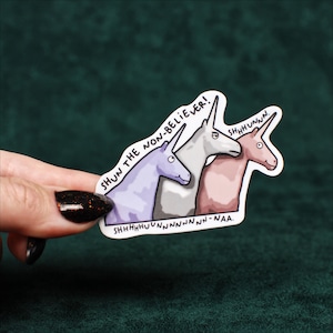 May include: A die-cut sticker featuring three cartoon unicorns in purple, gray, and pink. The text reads SHUN THE NON-BELIEVER! above the unicorns and SHHHHHUUUNNNNNNNNN-NAA. below them. The sticker has a white border and a matte finish.
