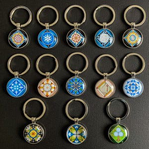 May include: Set of 15 silver keychains with round glass charms featuring colorful geometric designs. Each charm has a different pattern, including stars, flowers, and abstract shapes.