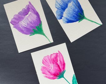 May include: Three rectangular cards featuring watercolor paintings of individual flowers. One card displays a purple flower, another a blue flower, and the third a pink flower. Each flower has a green stem and green leaves.