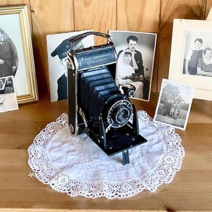 Könnte beinhalten: Eine schwarze Vintage-Faltkamera mit Balgenmechanismus und kreisförmiger Linseneinheit. Die Kamera hat ein Metallgehäuse mit einem Tragegriff und steht auf einer weißen, runden Spitzendeckchen. Das Objektivgehäuse trägt den Schriftzug Voigtlander.