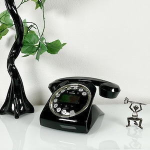 Könnte beinhalten: Ein schwarzes Festnetztelefon mit kreisförmigem Tastenfeld und digitalem Display. Das Gerät hat eine glänzende Oberfläche, einen oben aufliegenden Hörer und silberfarbene Akzente um den Wählbereich. Der Markenname Grundig ist zu sehen.