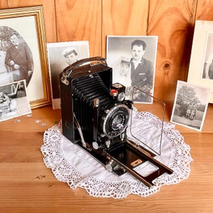 May include: A vintage folding bellows camera with a black leather-textured body, a silver lens assembly, and a metal frame viewfinder. The camera features a top handle, a collapsible bellows mechanism, and a front-mounted lens with visible text.