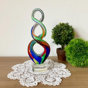 Peut inclure: Une sculpture décorative en verre coloré en forme de huit. La sculpture présente un mélange de couleurs vives, notamment le bleu, le rouge, le vert et l'orange, et est exposée sur une base ronde transparente. L'œuvre est posée sur une surface en bois avec un napperon en dentelle blanche.