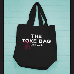 Könnte beinhalten: Schwarze Tragetasche mit dem Text "THE TOKE BAG" in Weiß, darunter "MARY JANE". Die Tasche hat eine rechteckige Form mit einer breiten Öffnung und zwei Henkeln. Der Hintergrund ist eine hellblaue Backsteinmauer.