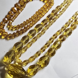 May include: Close-up of several strands of amber-coloured beaded necklaces. The beads vary in size and shape, with some being small and round, while others are larger and faceted. The necklaces are arranged on a white surface.