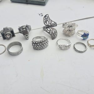Op de afbeelding: Een verzameling van elf zilverkleurige ringen met diverse ontwerpen, waaronder een ring in de vorm van een camera met een heldere steen, een filigraan halve maan ring, een cluster van kleine heldere steentjes en een blauwe sterring.