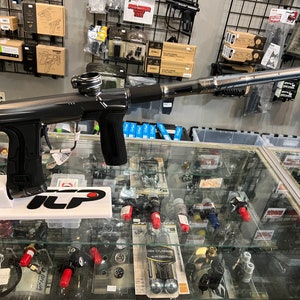 Può includere: Un marcatore da paintball nero con canna argentata e impugnatura nera. Il marcatore è su un espositore bianco con la scritta "ILP" stampata su di esso. Il marcatore è in una vetrina di vetro con altre attrezzature da paintball.
